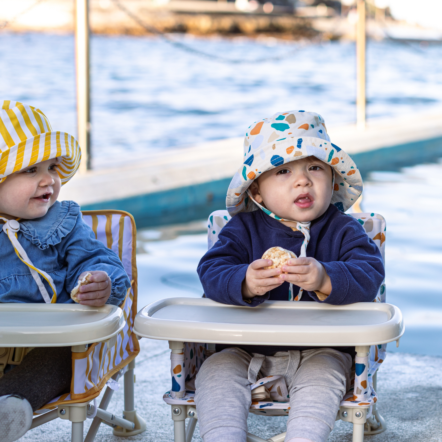 Two children sitting in high chairs by a body of water, eating.