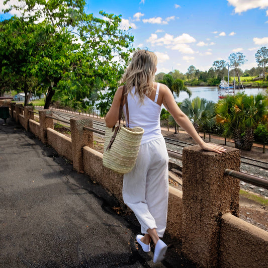 A woman seen from behind wearing a white tank top and white cropped pants, leaning on a stone bridge railing with a large woven straw tote bag over her shoulder, overlooking a scenic waterway with trees and a boat under a blue sky.