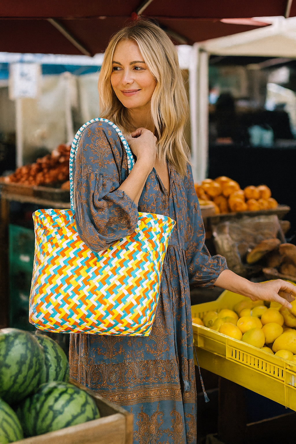 Woman holding a colorful patterned bag at an outdoor market with fruits and vegetables around.