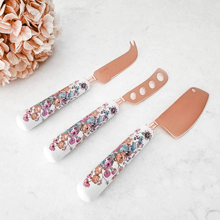 Set of three floral-patterned cheese knives on a white surface with a pink flower arrangement.