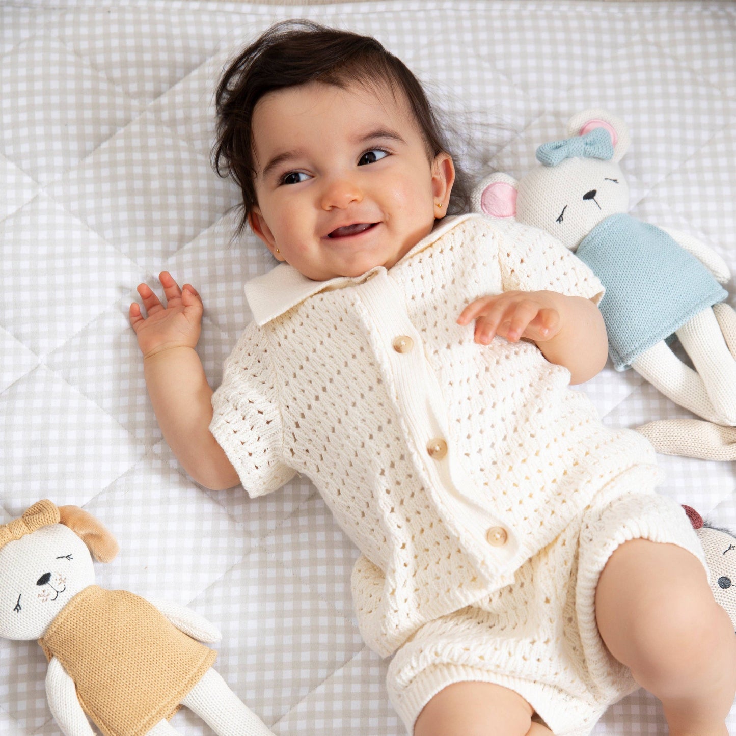 Baby in a white knitted outfit lying on a checkered blanket with toys.