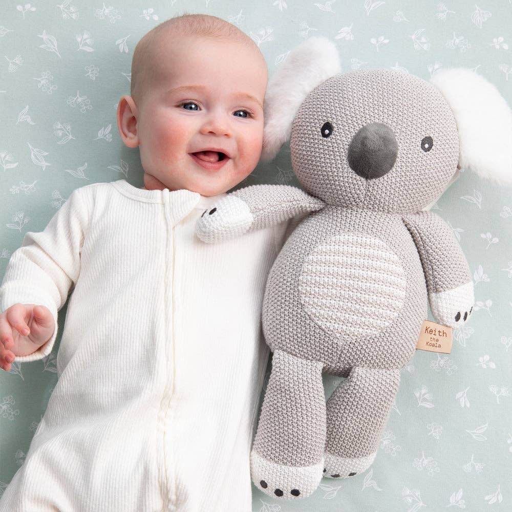 Baby in a white onesie holding a gray knitted Koala toy against a light blue background with floral patterns.