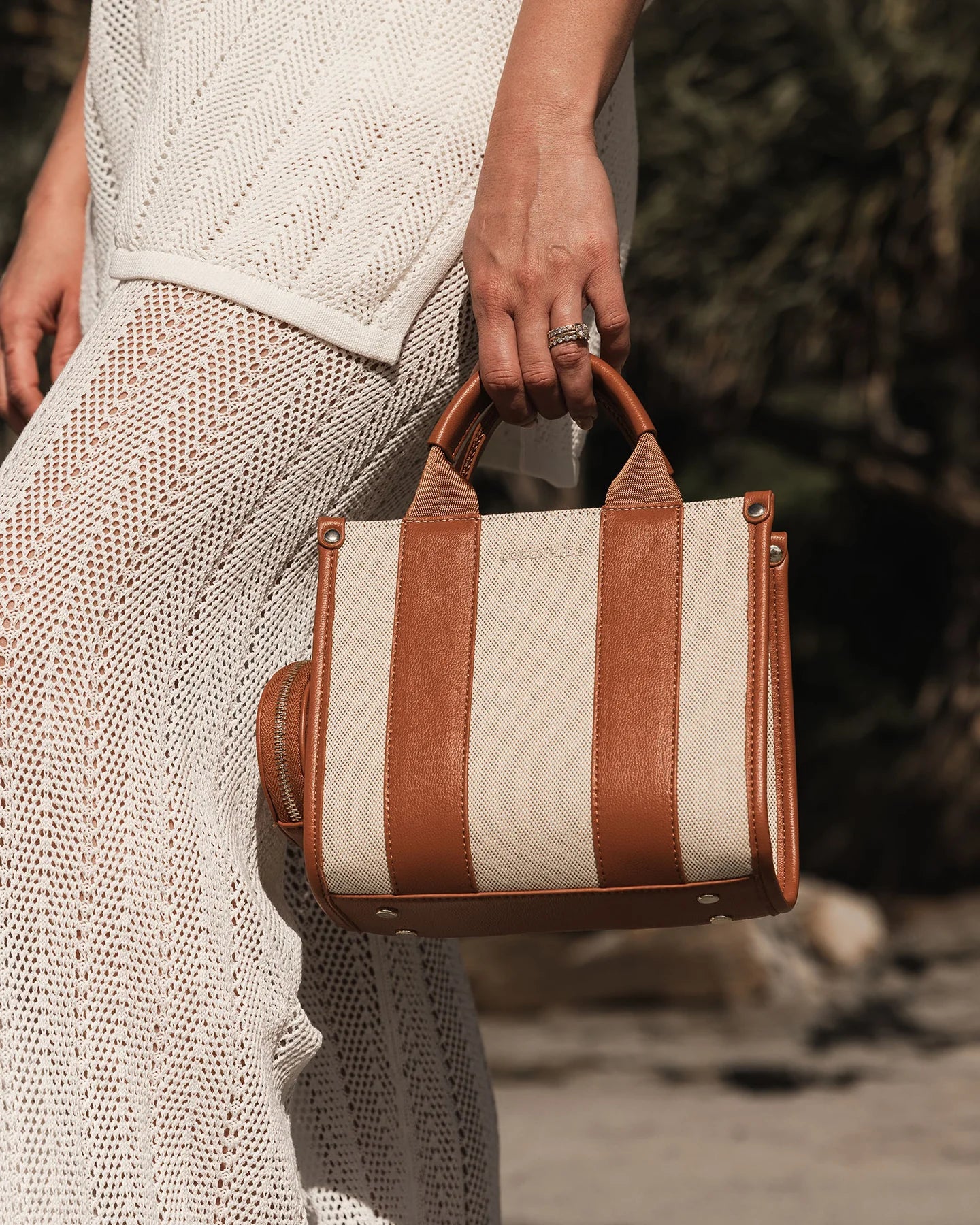 A close-up of a beige and brown tote bag with a shoulder strap, held by a person wearing a white crocheted dress.