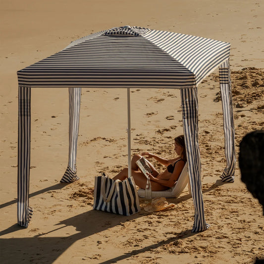 A woman relaxes under a black and white striped beach canopy on a sandy beach, with a matching striped tote bag beside her.