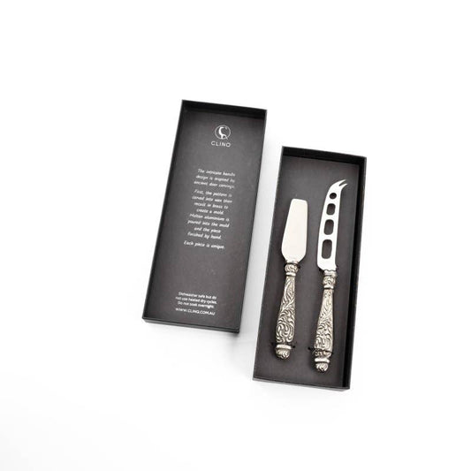 A cheese knife set with two knives featuring ornate handles, presented in a black box with a product description card.