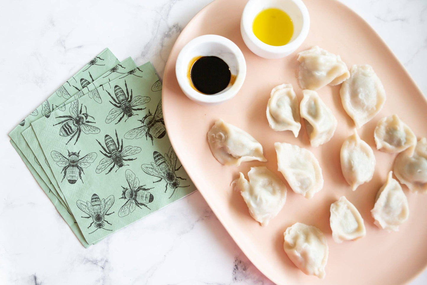 Dumplings on a pink plate with two small bowls of dipping sauce, accompanied by green napkins with bee designs.