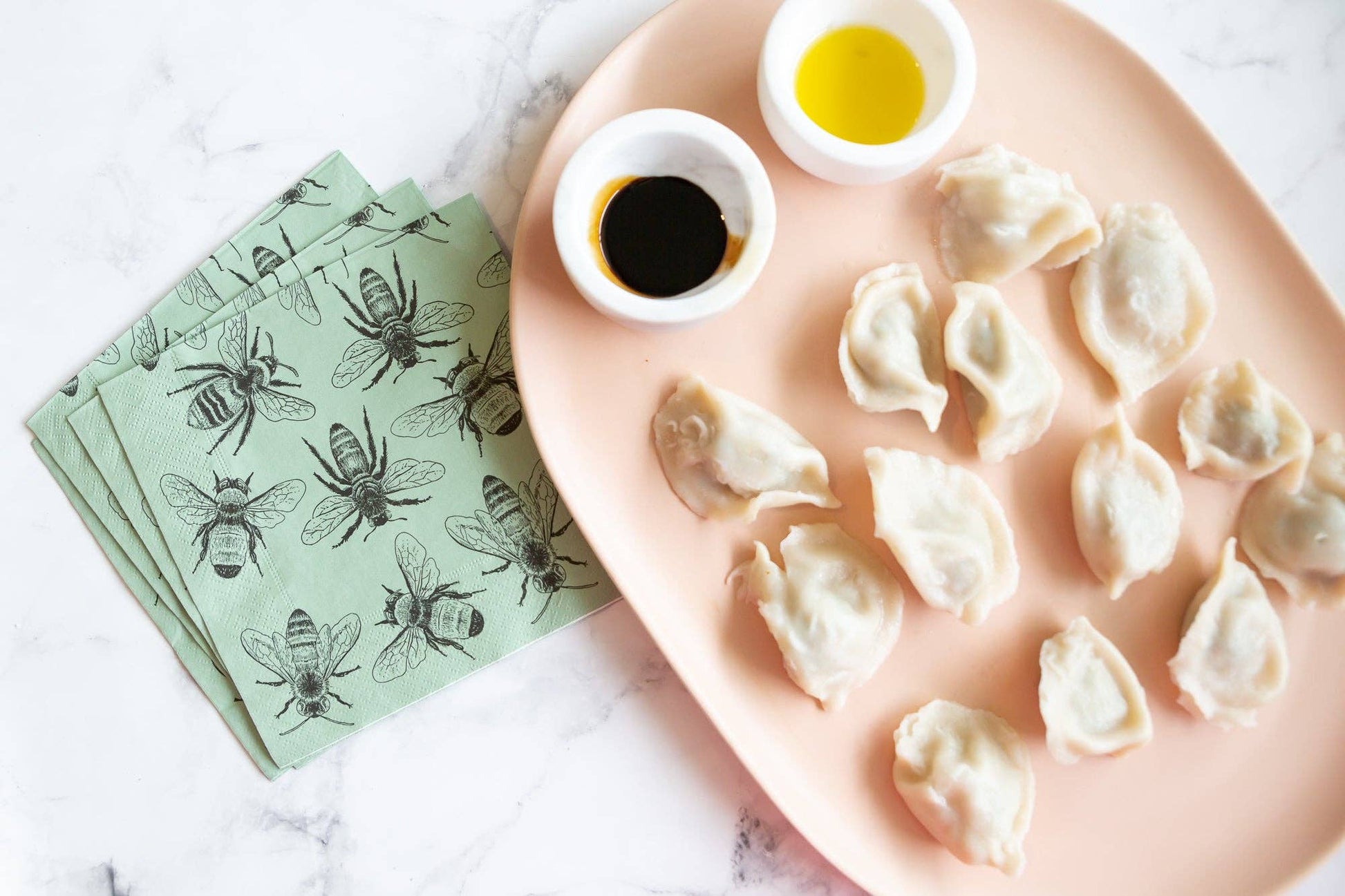 Dumplings on a pink plate with two small bowls of dipping sauce, accompanied by green napkins with bee designs.