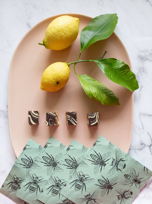 Two lemons on a branch with leaves on a beige plate, accompanied by brownies and a napkin with bee pattern.