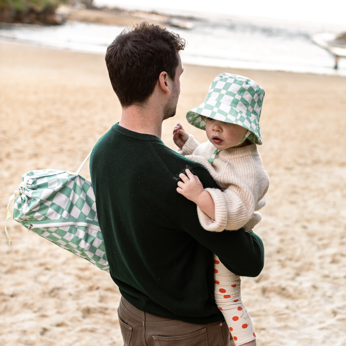 Man holding a child on a beach with a green checkered bag and matching bucket hat.