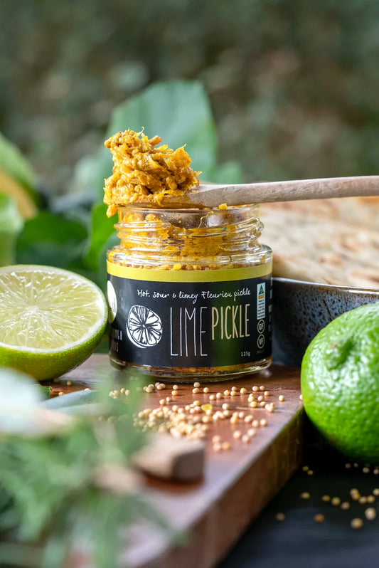 A jar of lime pickle with a wooden spoon lifting some of the pickled contents, styled with a halved lime, a whole lime, and scattered mustard seeds on a wooden board.