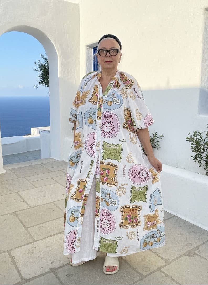 Woman wearing a patterned dress standing in a sunlit outdoor setting with white architecture and blue sea.