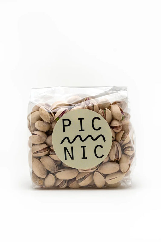 A clear plastic bag filled with pistachios, featuring a round beige label with the brand name 'PICNIC' in black text, set against a plain white background.