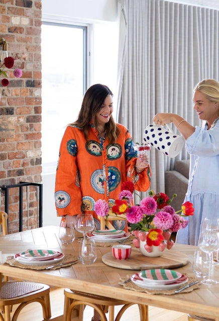 Two women setting a table with colorful plates and flowers in a bright room.