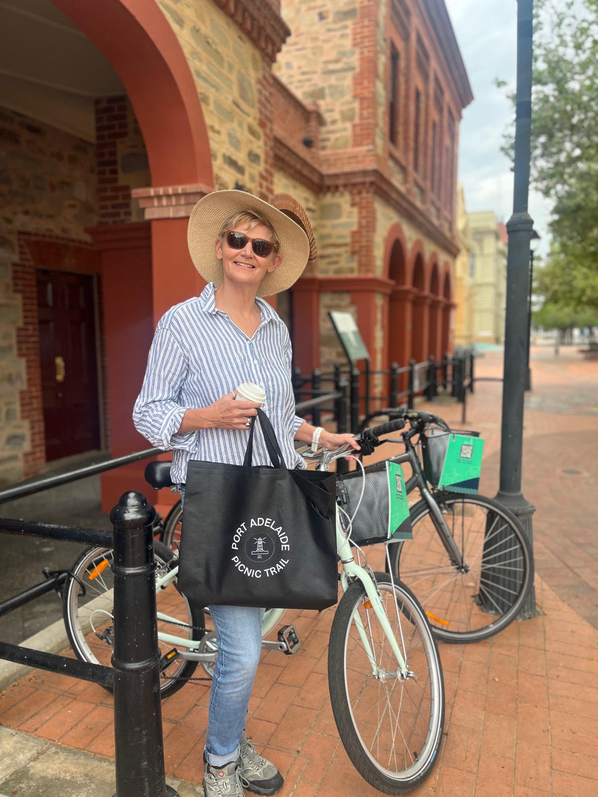 A woman wearing a straw hat and sunglasses stands on a brick sidewalk next to a bicycle, holding a white coffee cup and carrying a black tote bag with the text 'PORT ADELAIDE PICNIC TRAIL'.