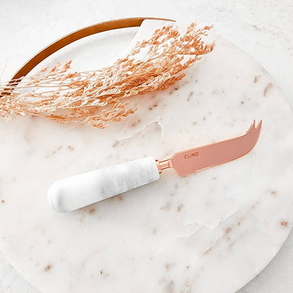 Rose gold and white cheese knife on a marble surface with dried grasses.