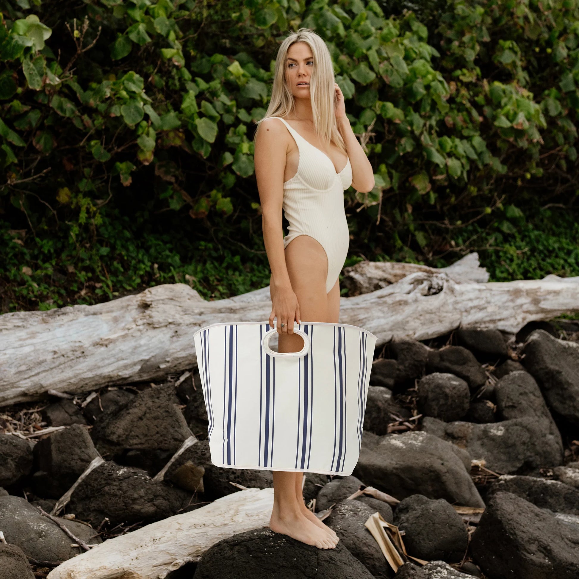 A person standing on rocks with a coastal blue and white striped carryall beach bag.