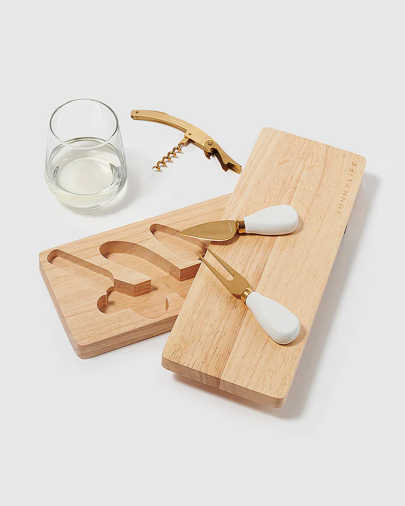 Wooden cheese board set with two knives, a corkscrew, and a glass of white wine on a light background.