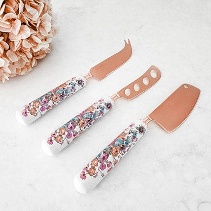Set of three floral-patterned cheese knives on a white surface with a pink flower arrangement.