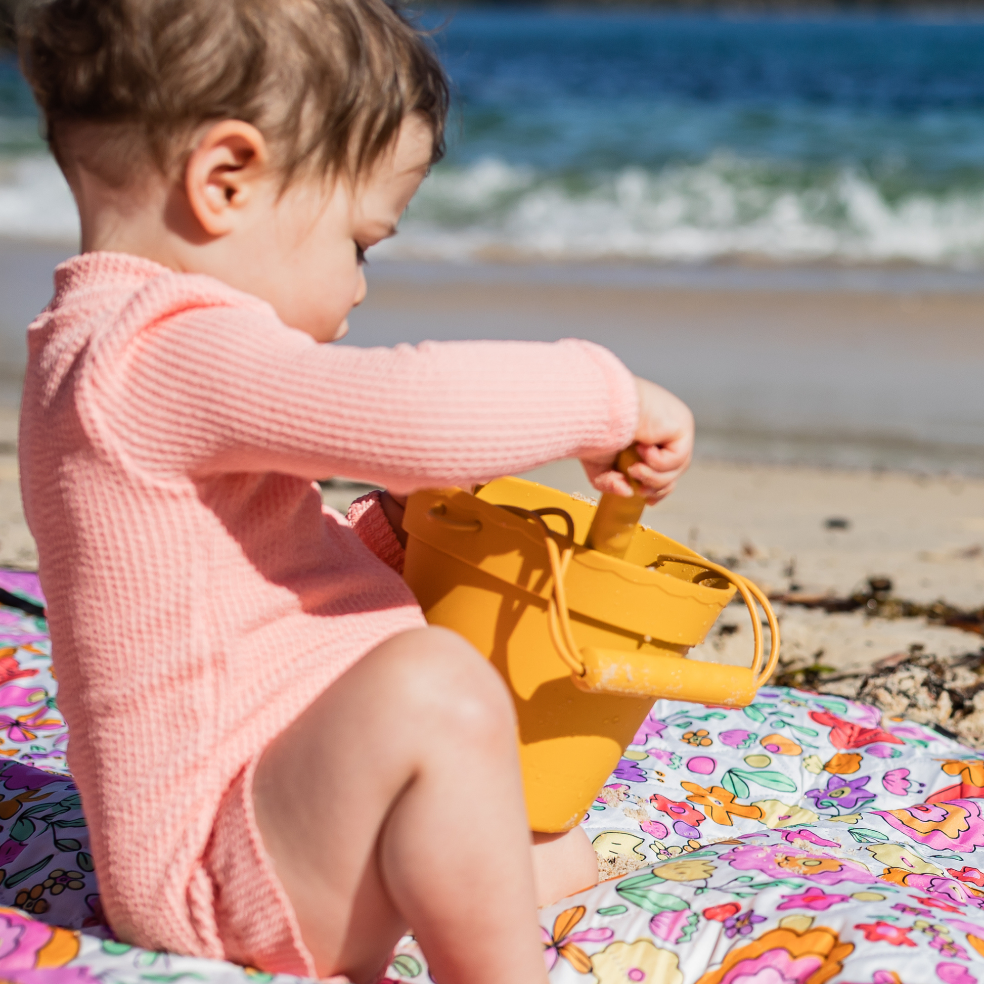 Child playing with a yellow sand bucket on a colorful beach mat by the ocean.