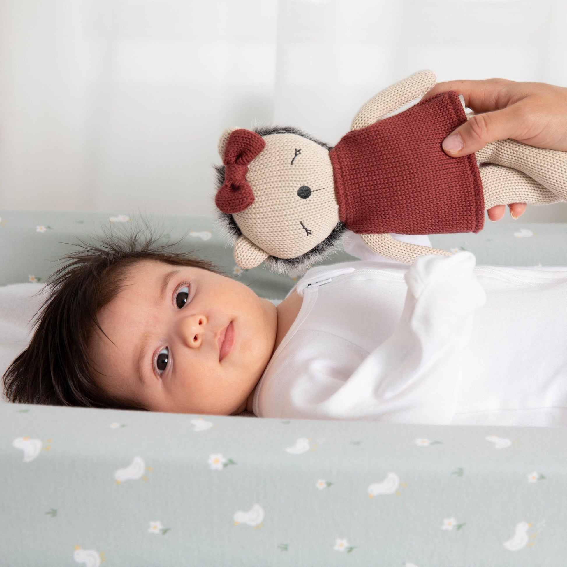 Baby lying on a crib with a hand holding a knitted toy