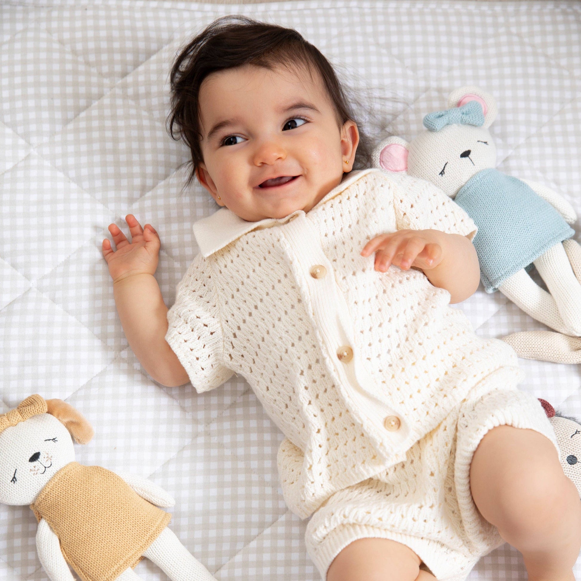 Baby in a white knitted outfit lying on a checkered blanket with toys.