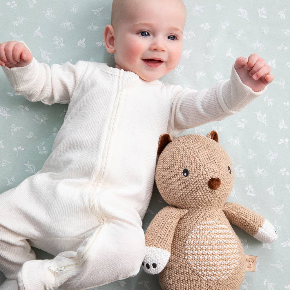 Baby in a white outfit sitting next to a brown knitted bear toy on a light blue background with floral patterns.