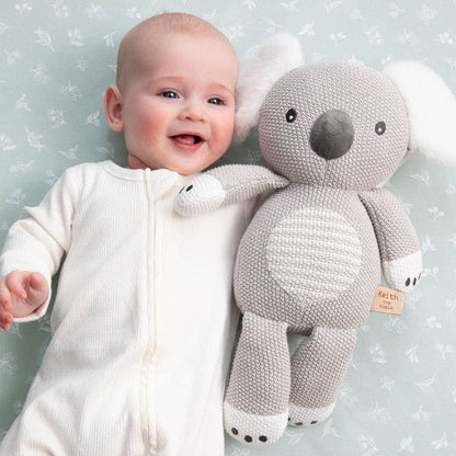 Baby in a white onesie holding a gray knitted Koala toy against a light blue background with floral patterns.