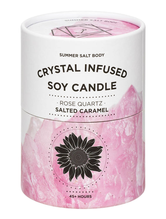 A cylindrical white and pink candle tin labeled 'CRYSTAL INFUSED SOY CANDLE' by Summer Salt Body, with rose quartz and salted caramel scent text and a black sunflower graphic on a white background.