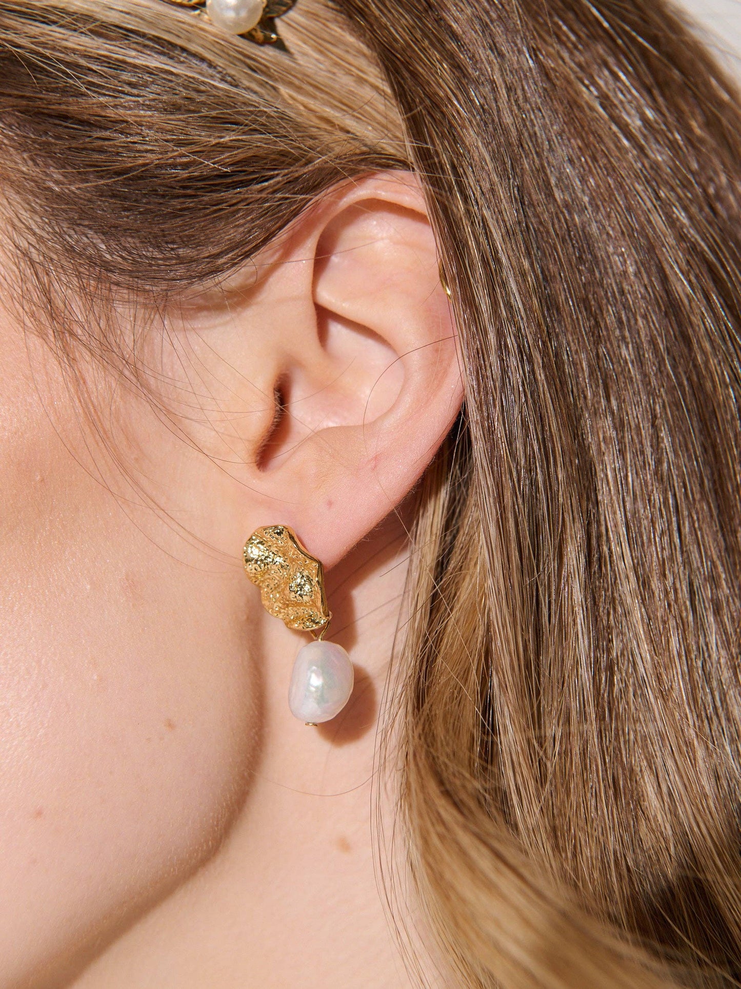 Close-up of an ear wearing a gold and pearl earring with blurred hair and skin in the background.