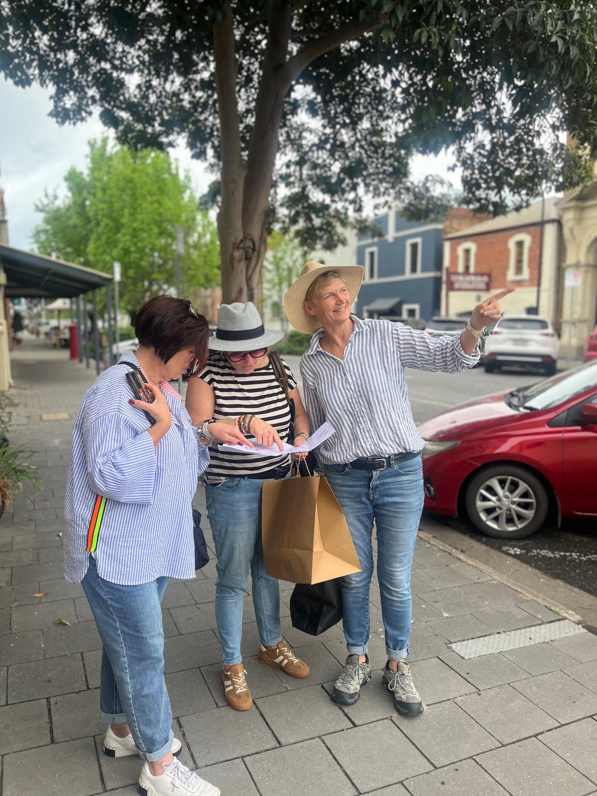 Three people standing on a city sidewalk under a tree, looking at a map or paper while one person points toward buildings in the background. A red car is parked on the street beside them.