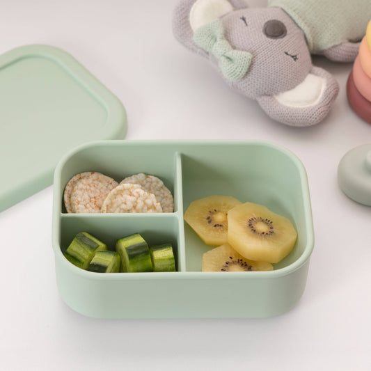 Green bento box with compartments containing rice cakes, kiwi slices, and green vegetables on a light surface.