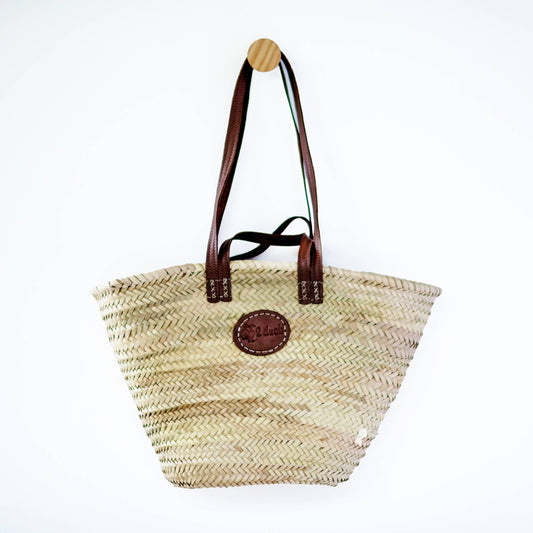 A natural woven straw tote bag with dark brown leather handles and a circular brown leather logo patch on the front, hanging from a wooden peg against a plain white background.