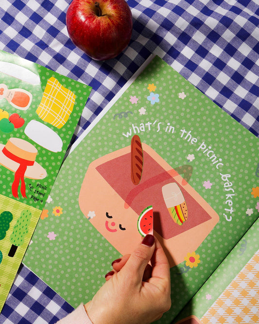 Children's book titled 'What's in the Picnic Basket?' on a checkered tablecloth with an apple beside it.