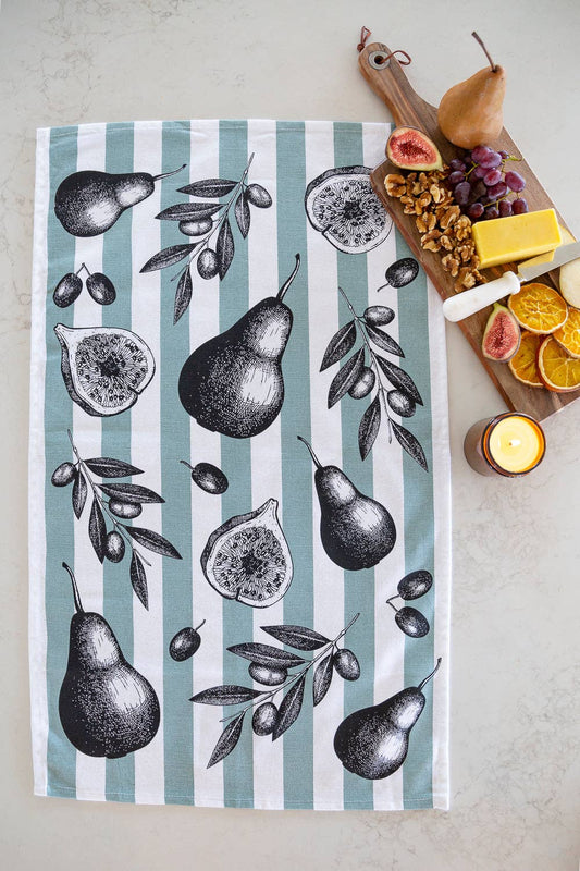 Tea towel with pear and fig design on a striped background, placed next to a cutting board with food.