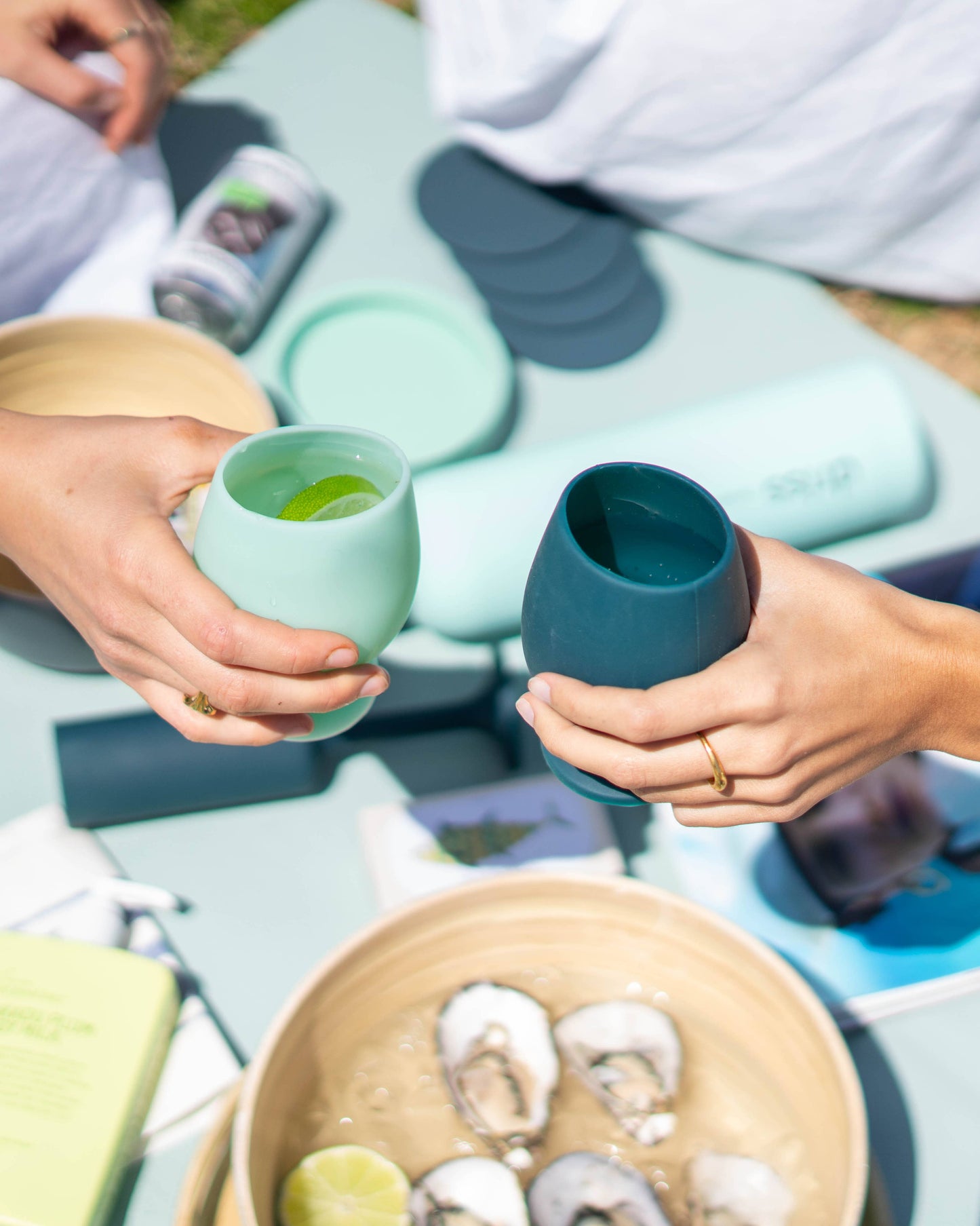 Two people holding Teal wines cups outdoors, with a bowl of snacks on a table.