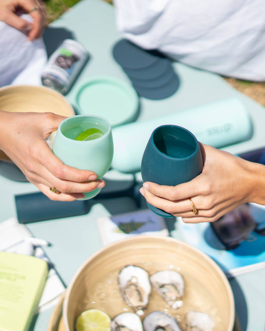 Two people holding Teal wines cups outdoors, with a bowl of snacks on a table.